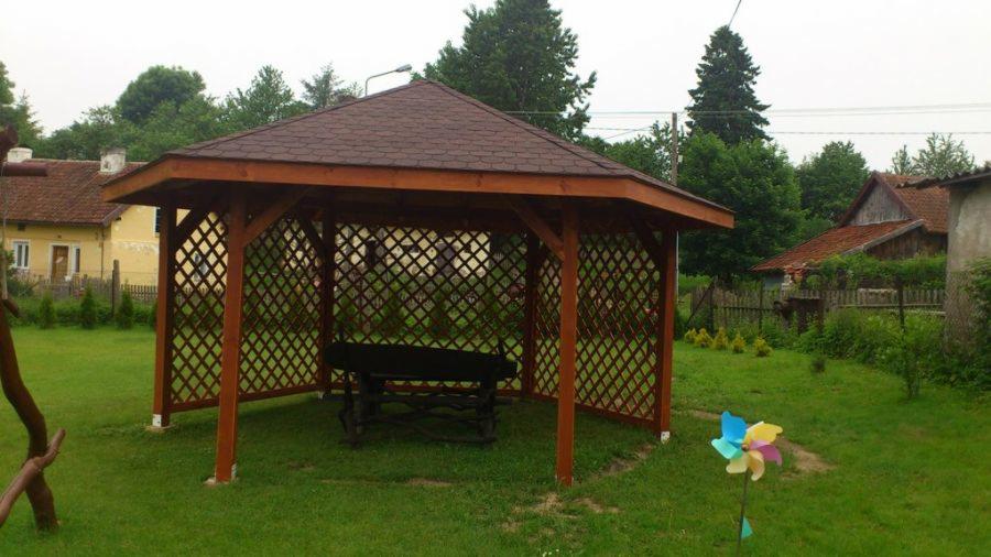 Drewniana altana ogrodowa z brązowym dachem bitumicznym, z ażurowymi ścianami i stołem wewnątrz, na tle zielonego trawnika i zabudowań wiejskich.