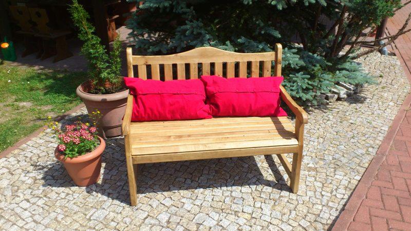 Drewniana ławka ogrodowa z dwoma czerwonymi poduszkami, ustawiona na brukowanej ścieżce, z kwiatami w doniczkach i iglakami w tle.