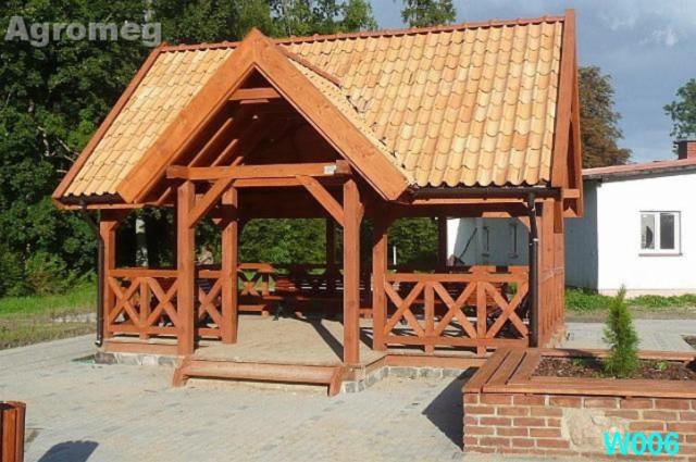 Drewniana altana z zadaszeniem z dachówki ceramicznej, widoczne elementy konstrukcyjne, balustrady z motywem krzyża, schody wejściowe i rabata z cegły klinkierowej, w tle budynek gospodarczy.