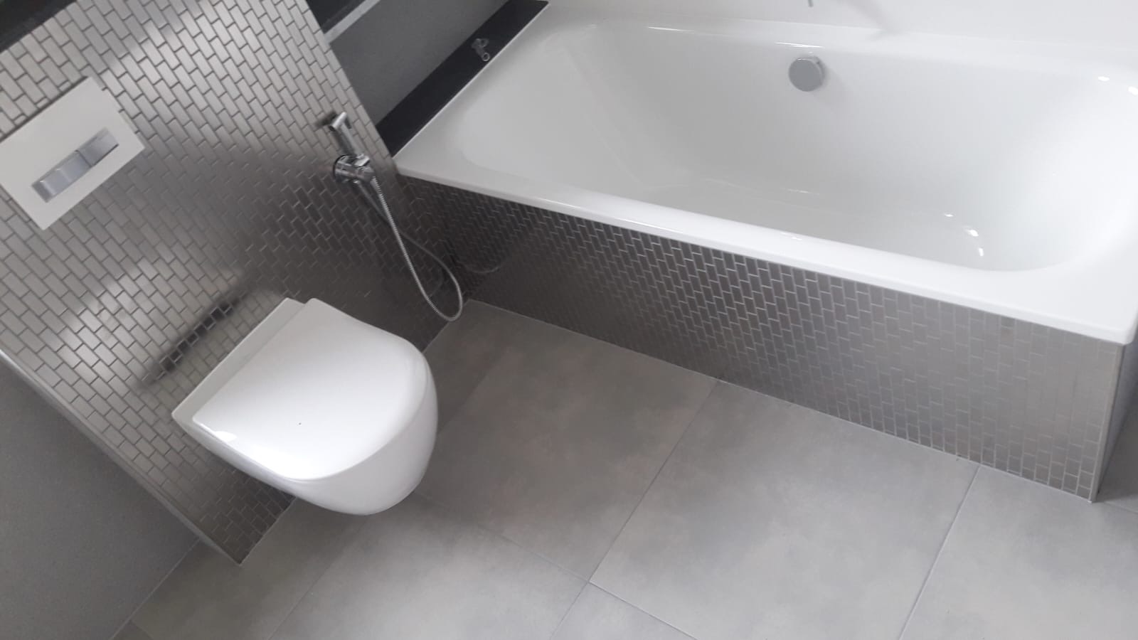 Nowoczesna łazienka z wanną obudowaną mozaiką w odcieniach szarości, wiszącą miską WC i szarą podłogą z płytek wielkoformatowych.