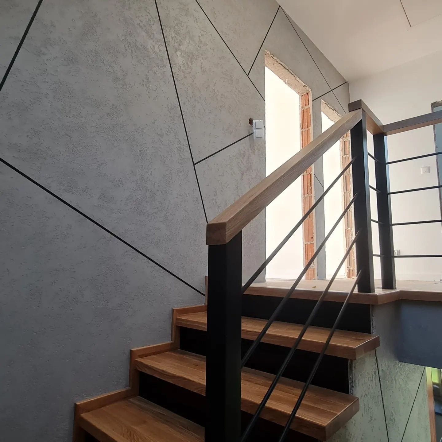 Klatka schodowa z drewnianymi stopniami i czarną metalową balustradą, ściana pokryta szarą dekoracyjną farbą z czarnymi geometrycznymi liniami, widok na otwarte przejście.