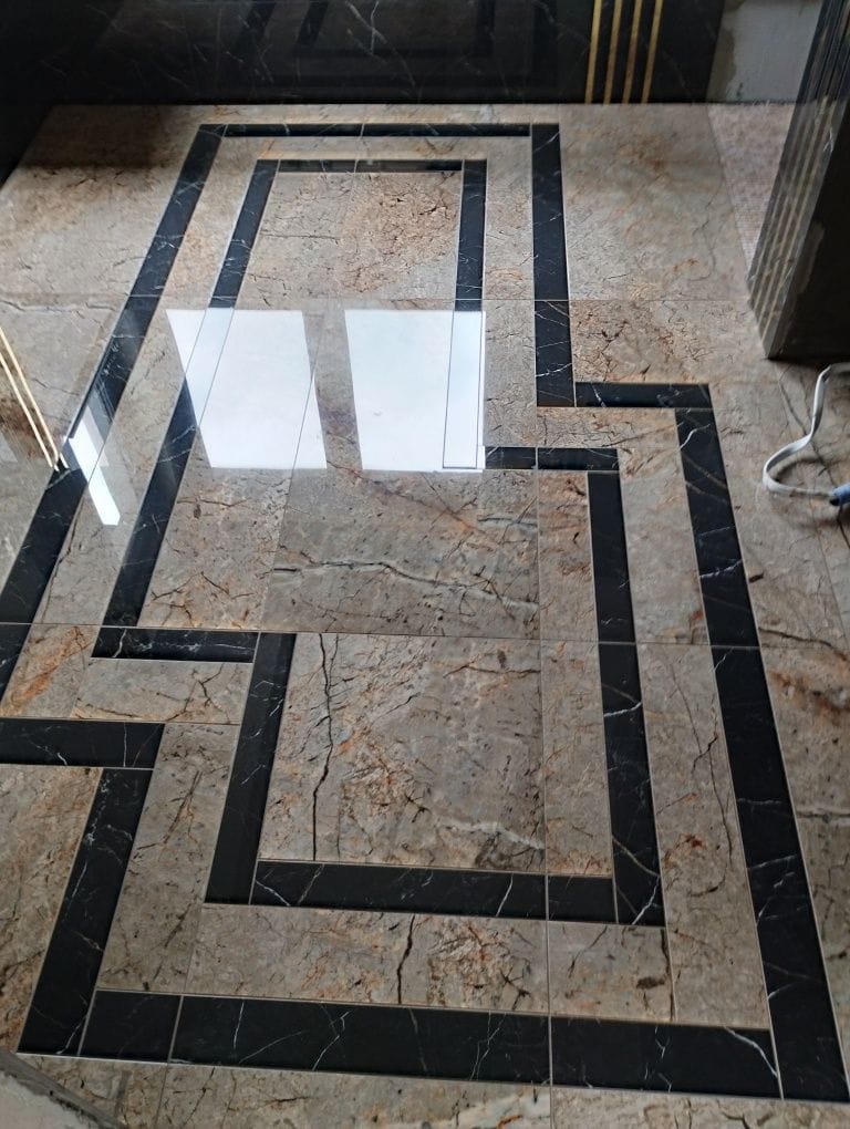 Ułożona podłoga z płytek w odcieniach beżu i brązu z czarnymi, geometrycznymi wstawkami tworzącymi wzór prostokątów. Na powierzchni widoczne odbicia światła.