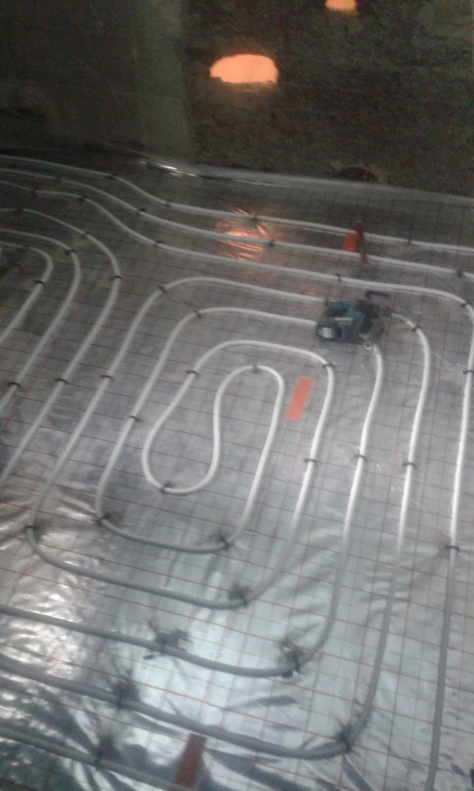 Rury ogrzewania podłogowego ułożone na folii z siatką, widoczne mocowania i narzędzie do ich montażu, surowy stan wykończenia.