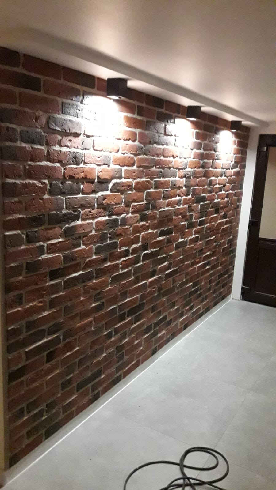 Ściana wyłożona dekoracyjną cegłą w odcieniach czerwieni i brązu, oświetlona czterema punktowymi lampami sufitowymi, z widocznym fragmentem szarej podłogi i zwisającym kablem.