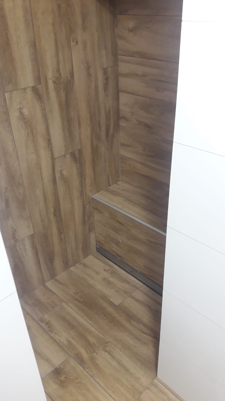 Ukończona wnęka z płytek imitujących drewno, z widoczną szyną prowadnicy drzwi przesuwnych i fragmentem białych drzwi.