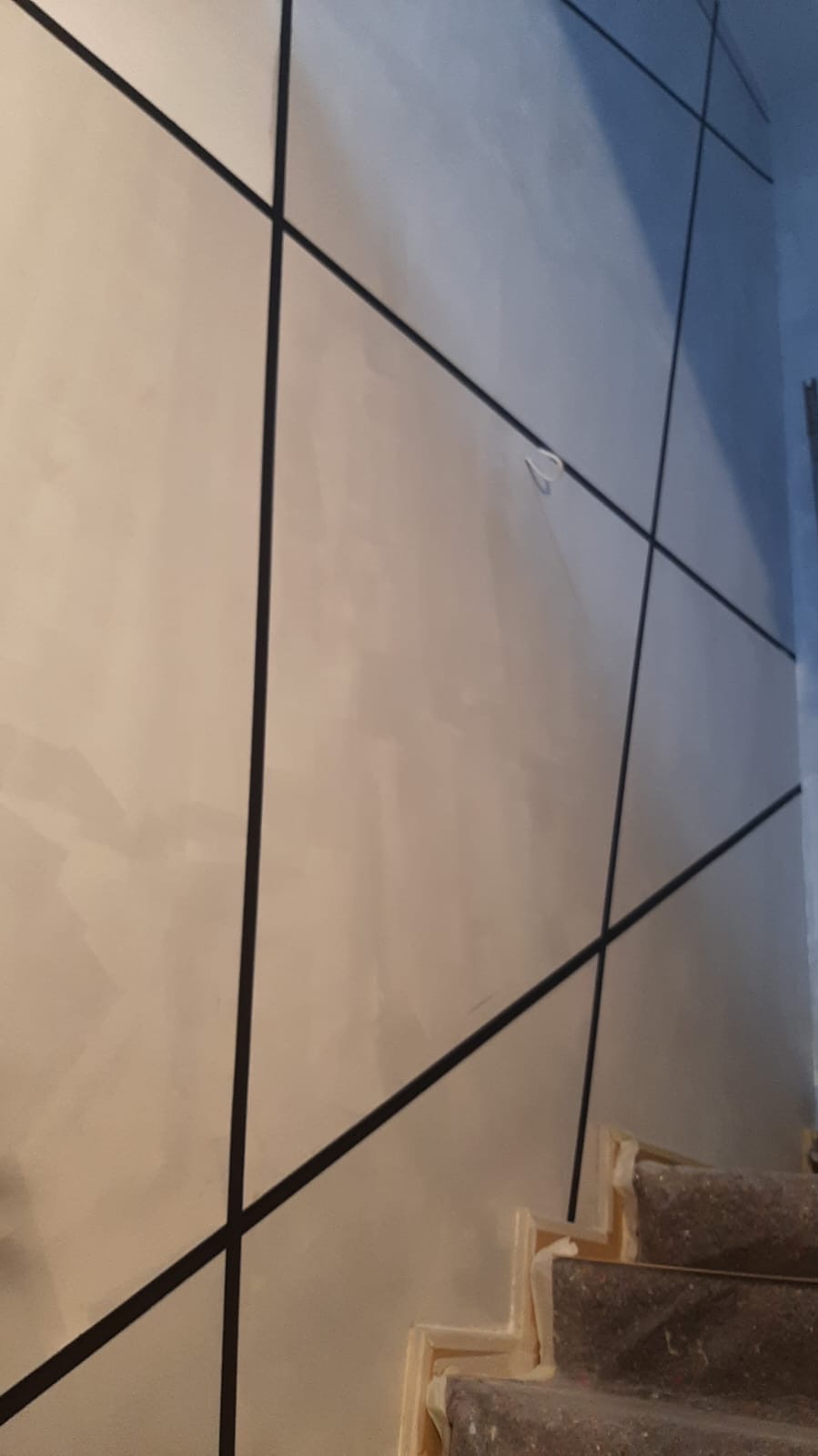 Ściana pomalowana na jasny kolor z czarnymi, ukośnymi liniami tworzącymi geometryczny wzór, widoczne schody z zabezpieczeniami.