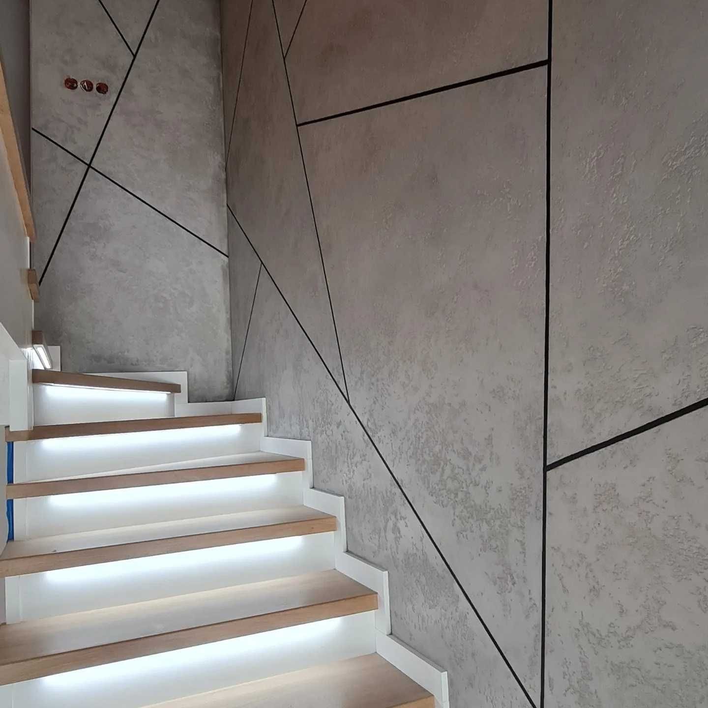 Ściana z dekoracyjnym tynkiem w odcieniach szarości, zdobiona czarnymi liniami tworzącymi geometryczny wzór, sąsiadująca z drewnianymi schodami z białymi podstopnicami i podświetleniem LED.