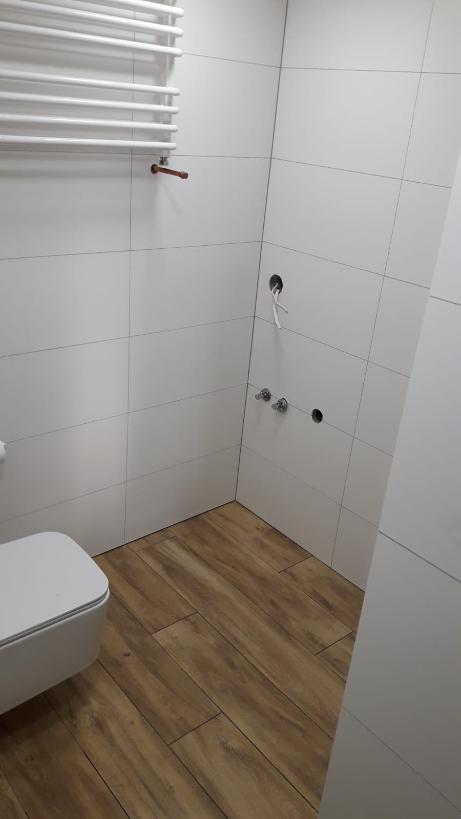 Wnętrze łazienki w trakcie remontu: białe, kwadratowe płytki na ścianach, drewnopodobna podłoga, widoczne otwory na baterie i wystające kable elektryczne, grzejnik drabinkowy.