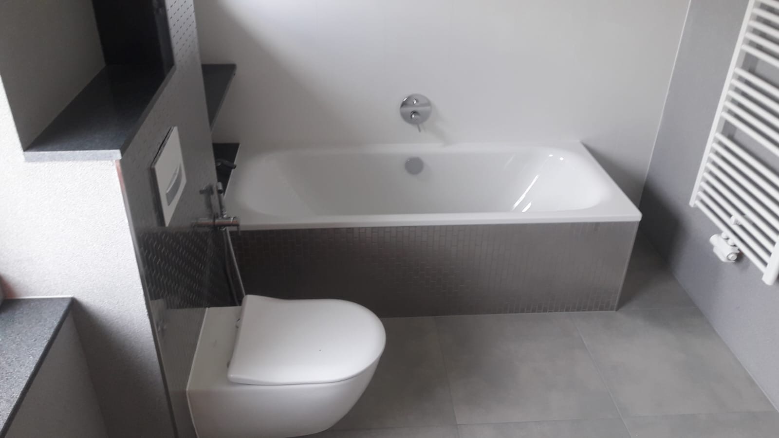 Nowoczesna łazienka w odcieniach szarości z wanną obłożoną mozaiką, podwieszaną toaletą i grzejnikiem drabinkowym.