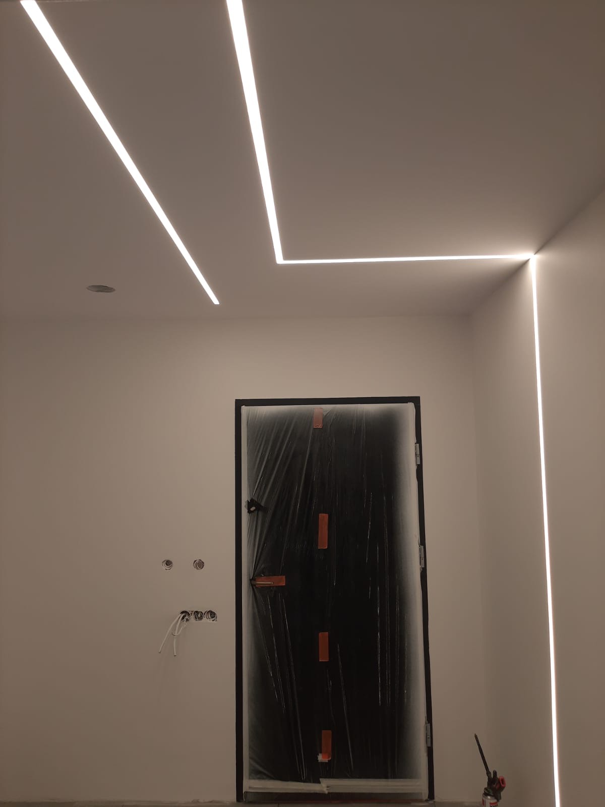 Nowoczesne oświetlenie sufitowe LED w kształcie litery L, czarne drzwi zabezpieczone folią malarską, widoczne kable elektryczne w ścianie