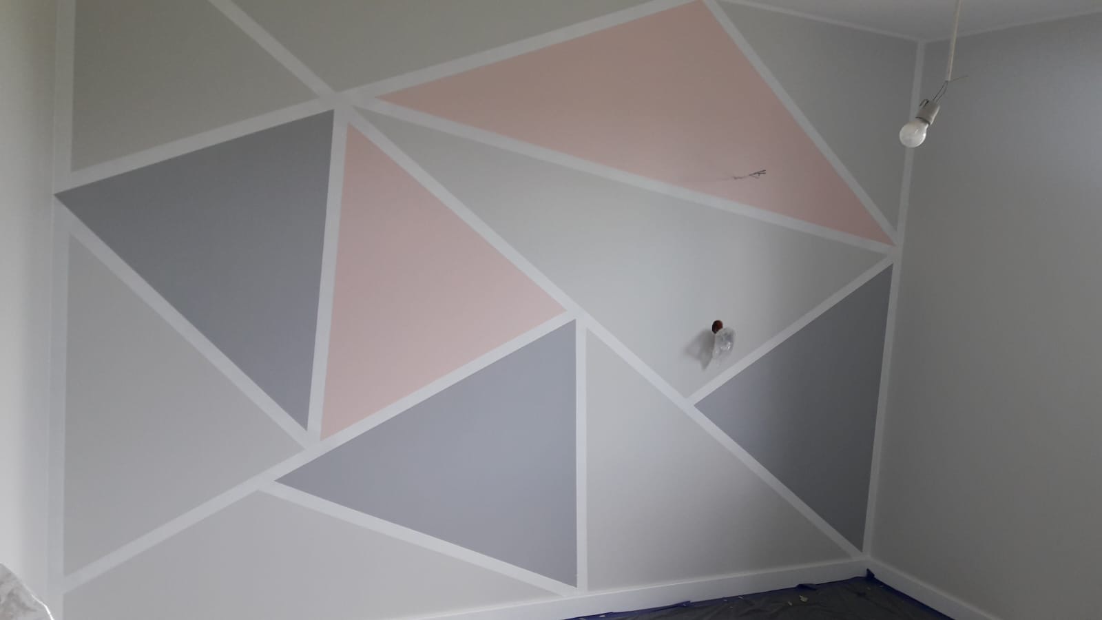 Ściana w pokoju pomalowana w geometryczne wzory z użyciem szarego, różowego i białego koloru, z widocznym gniazdkiem elektrycznym i żarówką na kablu.