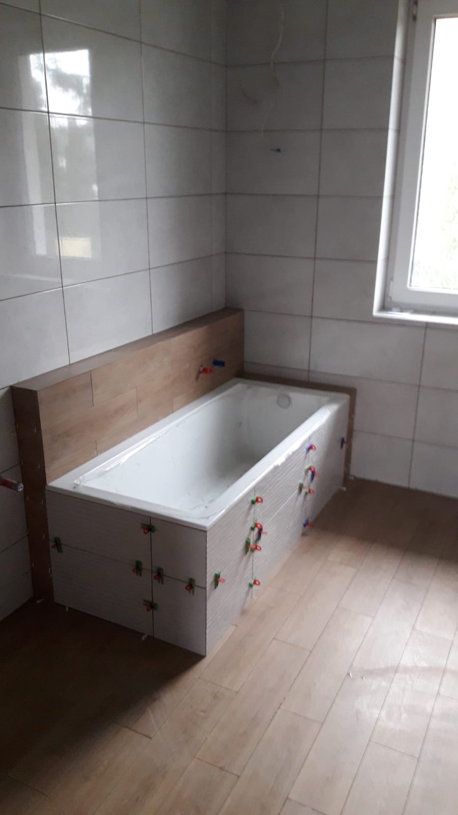 Biała wanna obudowana płytami gipsowymi, z widocznymi krzyżykami dystansowymi, na tle częściowo wyłożonej płytkami ściany i podłogi, podczas prac wykończeniowych w łazience.