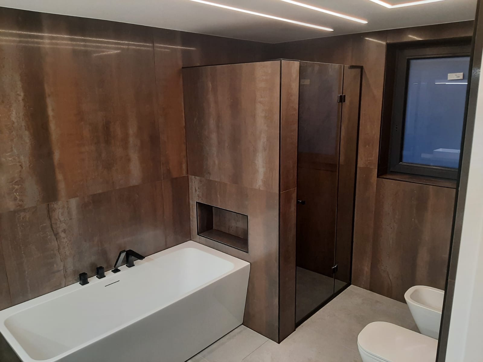 Nowoczesna łazienka z brązowymi płytkami imitującymi rdzę, białą wanną z czarną armaturą, kabiną prysznicową w czarnej ramie i podświetleniem LED na suficie.