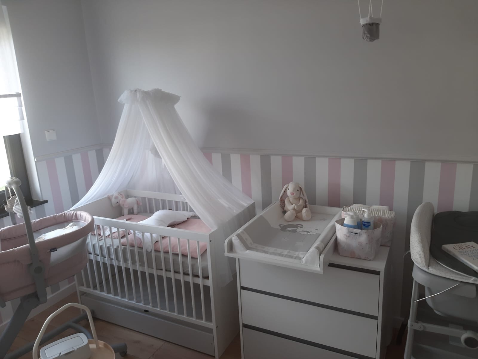 Pokój dziecięcy z białym łóżeczkiem z baldachimem, komodą z przewijakiem, tapetą w pastelowe pasy i pluszowym królikiem