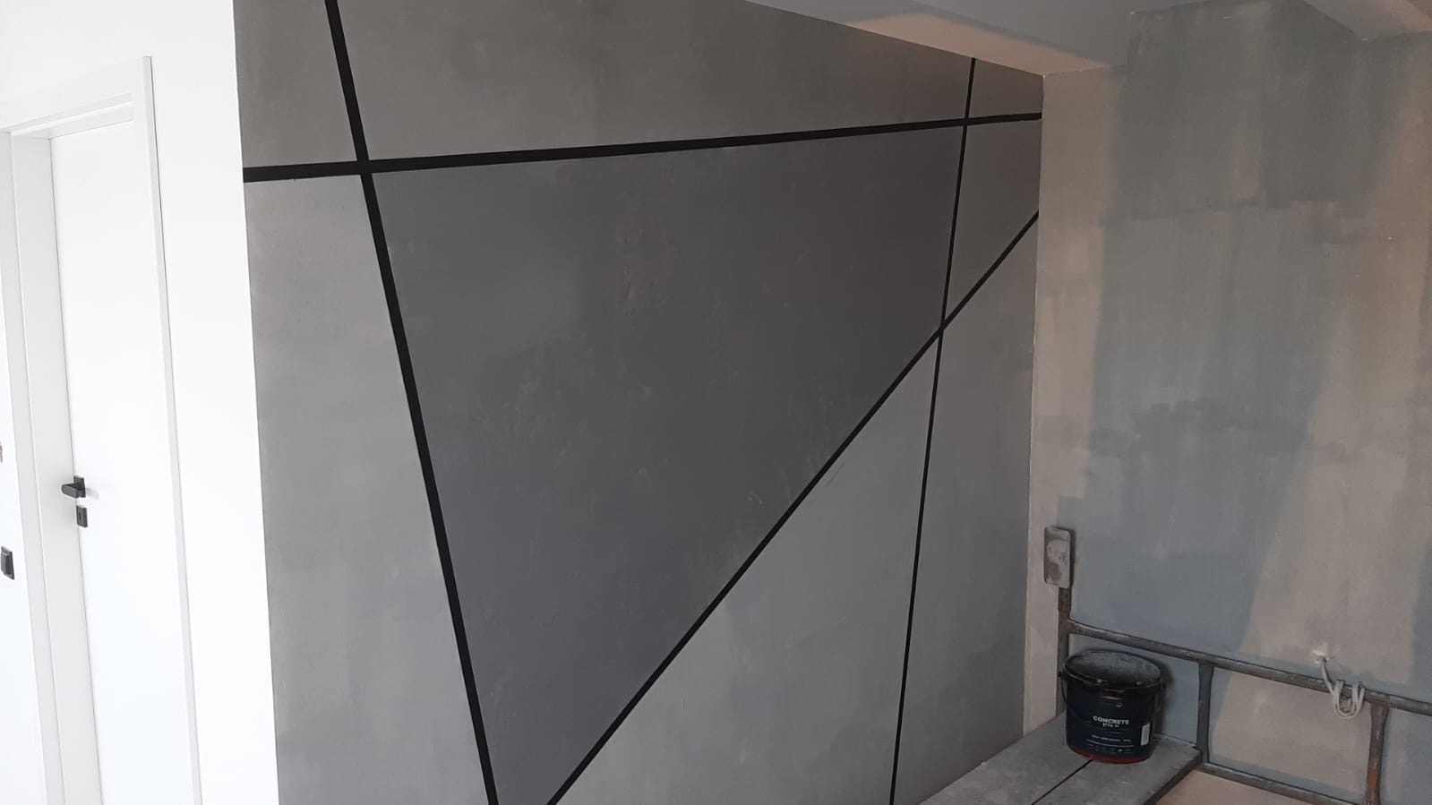 Ściana z szarą dekoracyjną fakturą i geometrycznymi czarnymi liniami, widoczna biała ościeżnica drzwi i puszka z farbą na rusztowaniu w tle.