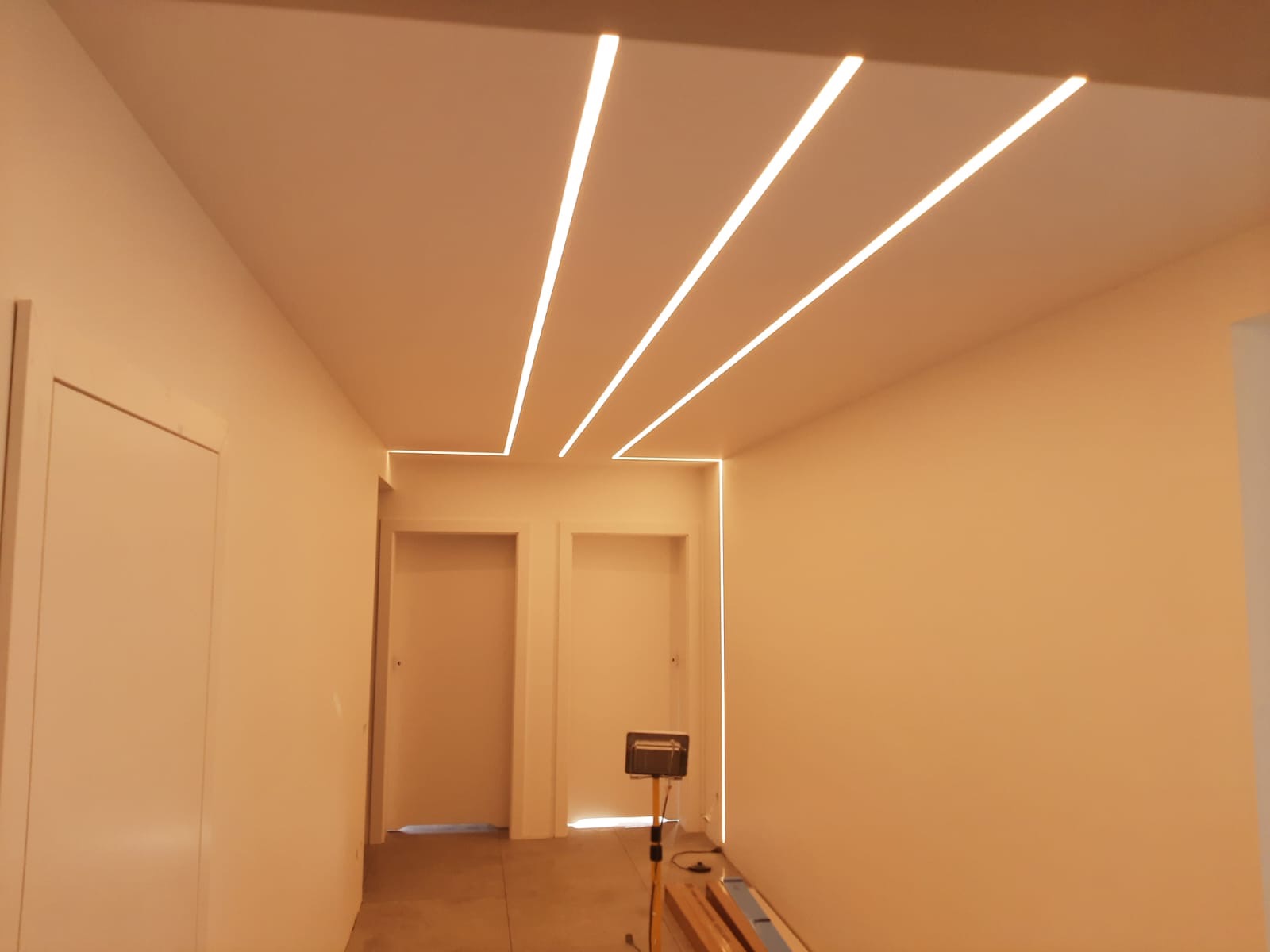 Korytarz z zamontowanym oświetleniem LED w suficie, tworzącym geometryczny wzór linii świetlnych, widoczna lampa robocza na statywie.