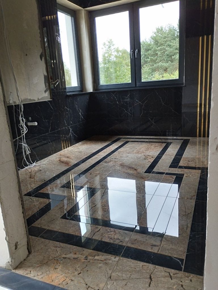 Łazienka w trakcie wykańczania, z marmurowymi płytkami podłogowymi w geometryczny wzór i czarnymi marmurowymi płytkami na ścianach, widoczne niedokończone instalacje elektryczne i szpachlowane ściany.