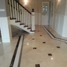 Jasna posadzka z kamienia naturalnego z ciemnymi, kwadratowymi intarsjami i dekoracyjnym obramowaniem, prowadząca do białych schodów z drewnianą balustradą w eleganckim wnętrzu z tapetą w subtelny...