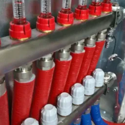 Rozdzielacz ogrzewania podłogowego z rotametrami i rurami w kolorach czerwonym i niebieskim, zainstalowany w Opolu. Widoczne metalowe elementy mocujące i izolacja termiczna na rurach.