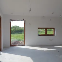 Surowe wnętrze domu w trakcie wykończenia, z oknami z brązowymi ramami, świeżo pomalowanymi na biało ścianami i surową podłogą, widoczne wystające kable od oświetlenia.