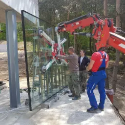 Montaż dużej szyby okiennej za pomocą przyssawki podnoszonej przez dźwig, widoczni trzej pracownicy w ubraniach roboczych na tle lasu.