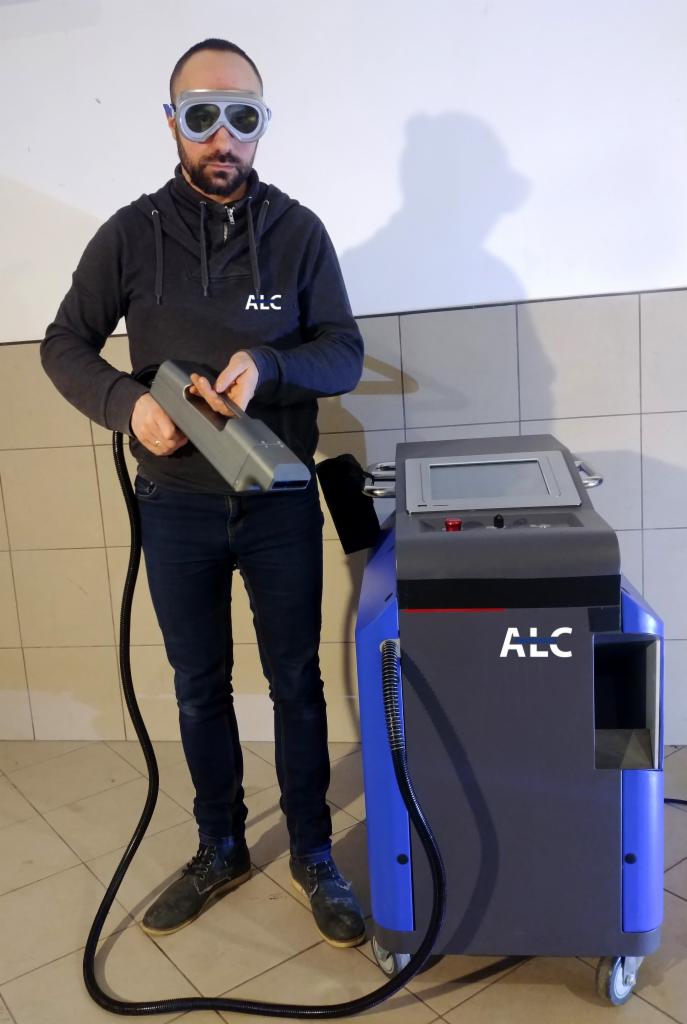 Mężczyzna w okularach ochronnych prezentuje urządzenie laserowe z logo 'ALC', prawdopodobnie do czyszczenia powierzchni, na tle ściany z płytek.
