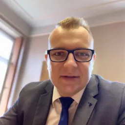 Portret mężczyzny w okularach i garniturze, z niebieskim krawatem, siedzącego przy biurku w jasnym biurze. Uśmiech, profesjonalny wygląd.