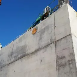 Wysoki, betonowy słup w trakcie budowy, z widocznymi elementami zbrojenia i śladami po szalunkach, na tle błękitnego nieba.