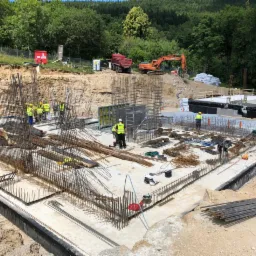 Budowa fundamentów: zbrojenie, szalunki i pracownicy w żółtych kamizelkach, w tle koparka i ciężarówka, teren budowy otoczony zielenią.