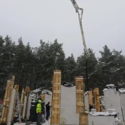 Wznoszenie ścian domu z bloczków betonowych w zimowej scenerii, widoczna pompa do betonu i pracownicy w odblaskowych kamizelkach.