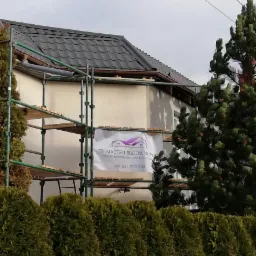 Dom w trakcie prac elewacyjnych, widoczne rusztowania, fragment ocieplonej ściany i baner firmy budowlanej z numerem telefonu.