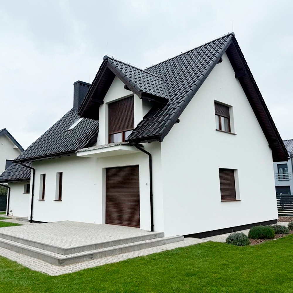 Biały dom jednorodzinny z ciemną dachówką i brązowymi roletami zewnętrznymi, widok z poziomu trawnika w pochmurny dzień. Nowoczesna architektura.