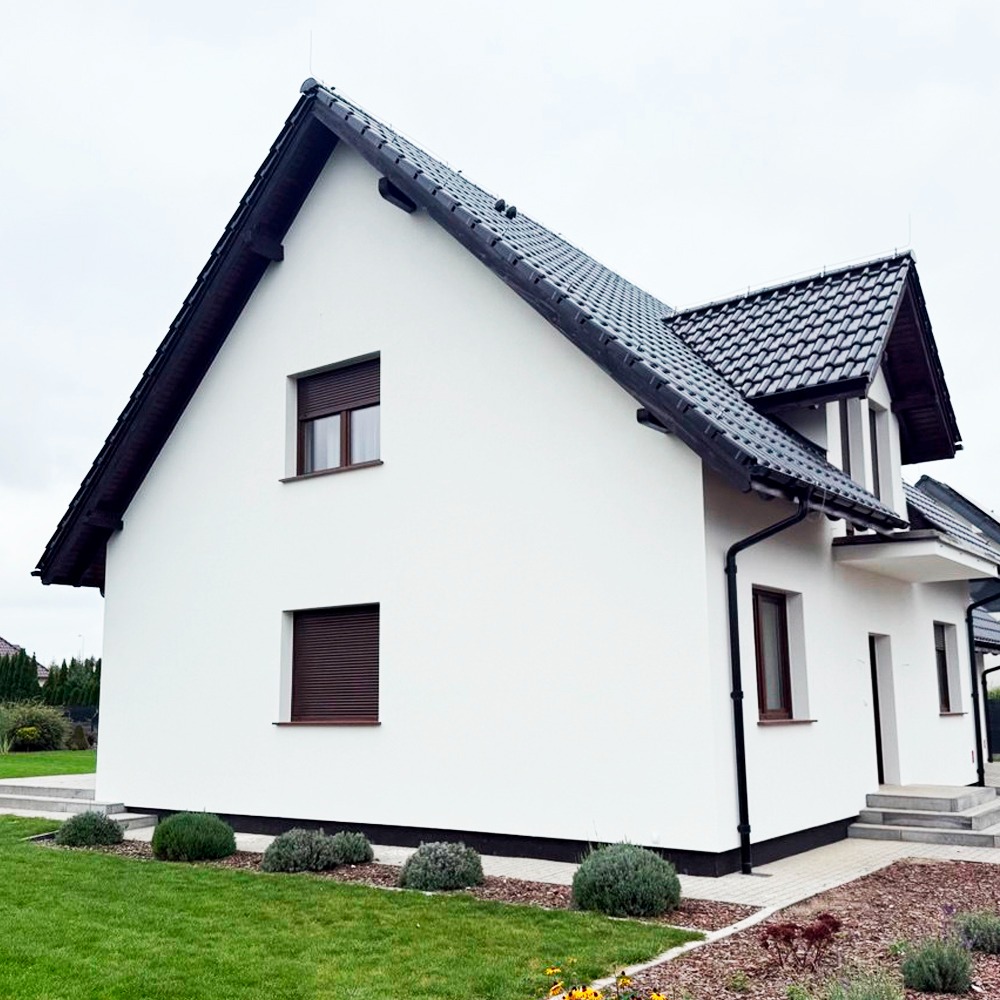 Biały dom jednorodzinny z ciemną dachówką i brązowymi roletami zewnętrznymi, widok z boku na zadbany trawnik i rabaty.