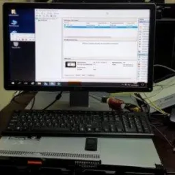 Monitor wyświetlający interfejs oprogramowania serwerowego, klawiatura spoczywająca na obudowie serwera rack, w tle monitory i kartony.