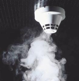 Biały czujnik dymu zamontowany na suficie z widocznym dymem unoszącym się w jego kierunku.