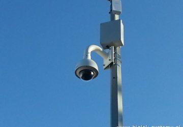 Kamera obrotowa PTZ zamontowana na metalowym słupie na tle błękitnego nieba. Widoczna puszka instalacyjna i antena. Ujęcie z dołu.