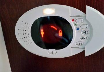 Panel sterowania z podświetlanymi ikonami: sylwetka osoby i zamknięta kłódka, sygnalizujące status bezpieczeństwa. Biała obudowa zamontowana na ciemnym, drewnianym tle.