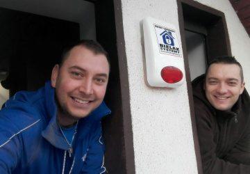 Uśmiechnięci mężczyźni, jeden w niebieskiej bluzie, drugi w brązowej kurtce, pozują w oknie obok białej obudowy alarmu z czerwonym światłem i logo firmy Bielak Systemy.