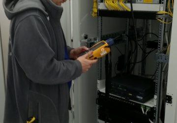 Technik w Gryfowie Śląskim sprawdza połączenia kablowe w szafie rack za pomocą miernika. Widoczne żółte kable i urządzenia sieciowe.
