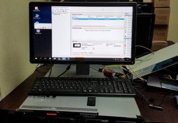 Monitor wyświetlający interfejs oprogramowania serwerowego, klawiatura spoczywająca na obudowie serwera rack, w tle monitory i kartony.