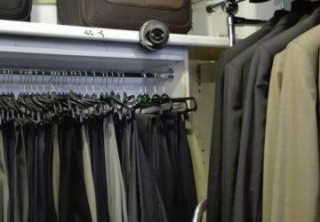 Kamera przemysłowa zamontowana na półce nad wieszakami z ubraniami w sklepie odzieżowym. Widoczne czarne i szare spodnie oraz beżowe marynarki. U góry brązowa torba.