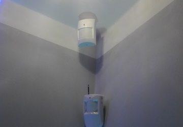 Dwa czujniki ruchu zamontowane w rogu pomieszczenia z szaro-białymi ścianami i jasnoniebieskim sufitem, widoczny cień jednego z czujników.