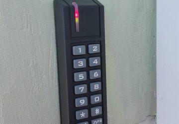 Czarny panel kontrolny dostępu z klawiaturą numeryczną i czerwoną diodą LED zamontowany na jasnej ścianie. Widoczne ślady użytkowania na ścianie w dolnym rogu.