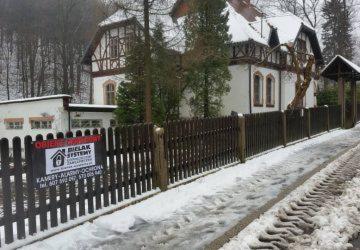 Tablica reklamowa systemu alarmowego na tle zasypanego śniegiem ogrodzenia posesji z domem w stylu szwajcarskim.