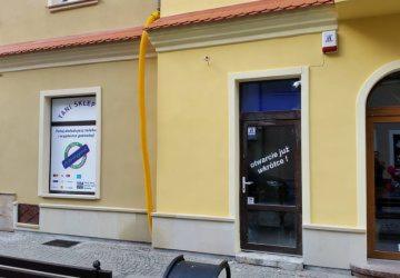 Zewnętrzny widok budynku z żółtą rurą spustową, oknem z reklamą 'Tani Sklep' i drzwiami z napisem 'otwarcie już wkrótce!', oraz zamontowanym alarmem na ścianie.