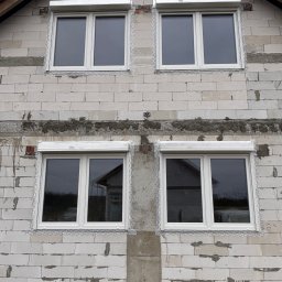 Demonti Karol Prybacki - Montaż białych okien z roletami zewnętrznymi w surowym budynku z białego bloczka. Widoczne zabezpieczenia okien folią. Dom w trakcie budowy.