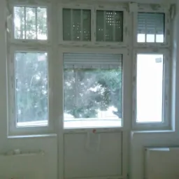 Wnętrze pokoju z zamontowanymi białymi oknami z roletami w różnych konfiguracjach, widoczny grzejnik i wiadro z narzędziami malarskimi.