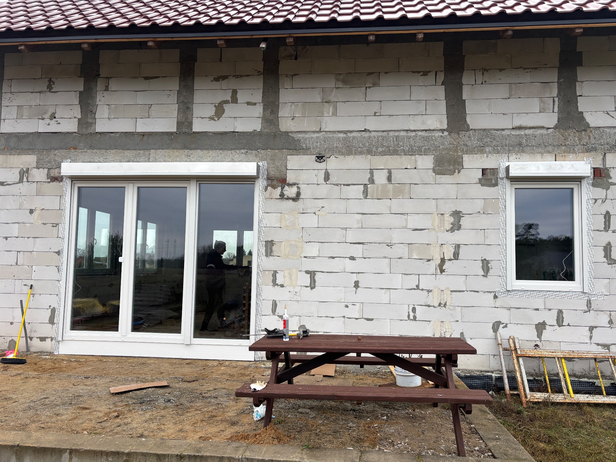 Montaż okien i rolet zewnętrznych w surowym budynku. Widoczne narzędzia pracy i drabina. Okno trzyskrzydłowe i pojedyncze z zamontowanymi roletami.