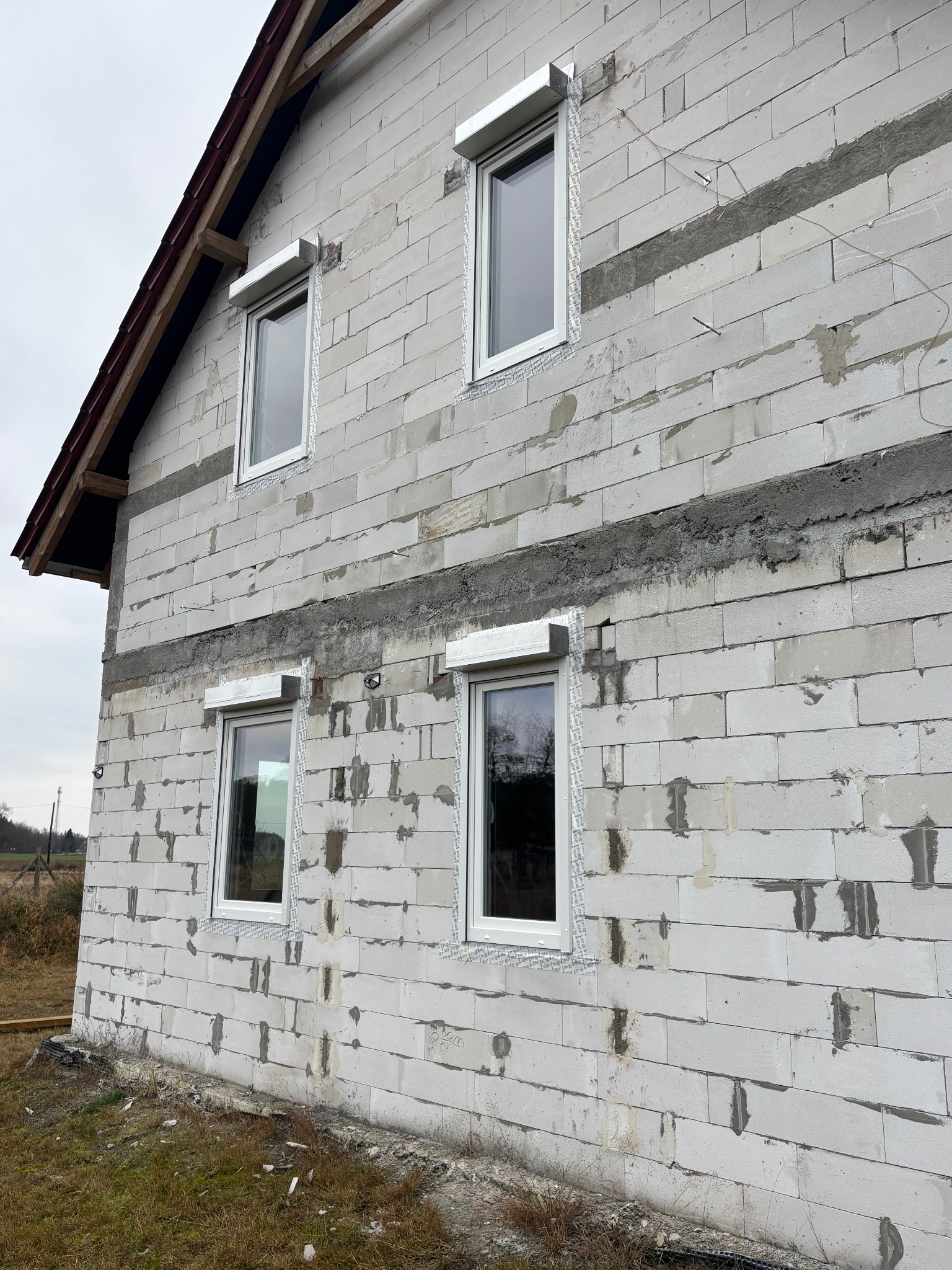 Surowy budynek z białego bloczka z zamontowanymi oknami i roletami zewnętrznymi w kolorze białym. Widoczne ślady zaprawy i niedokończone wykończenie.