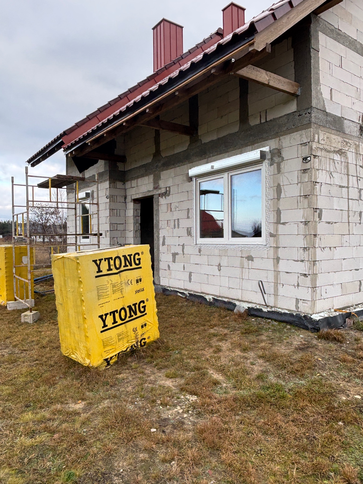 Budowa domu z białych bloczków Ytong, widoczne rusztowania i zamontowane okno z roletą zewnętrzną. Częściowo ocieplony budynek w trakcie prac wykończeniowych.